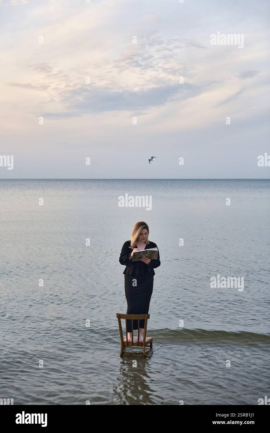 Una giovane donna adulta sta su una sedia di legno nelle calme acque della spiaggia, leggendo un libro. Il cielo sereno e le onde dolci creano un'atmosfera tranquilla ma surreale, simbo Foto Stock