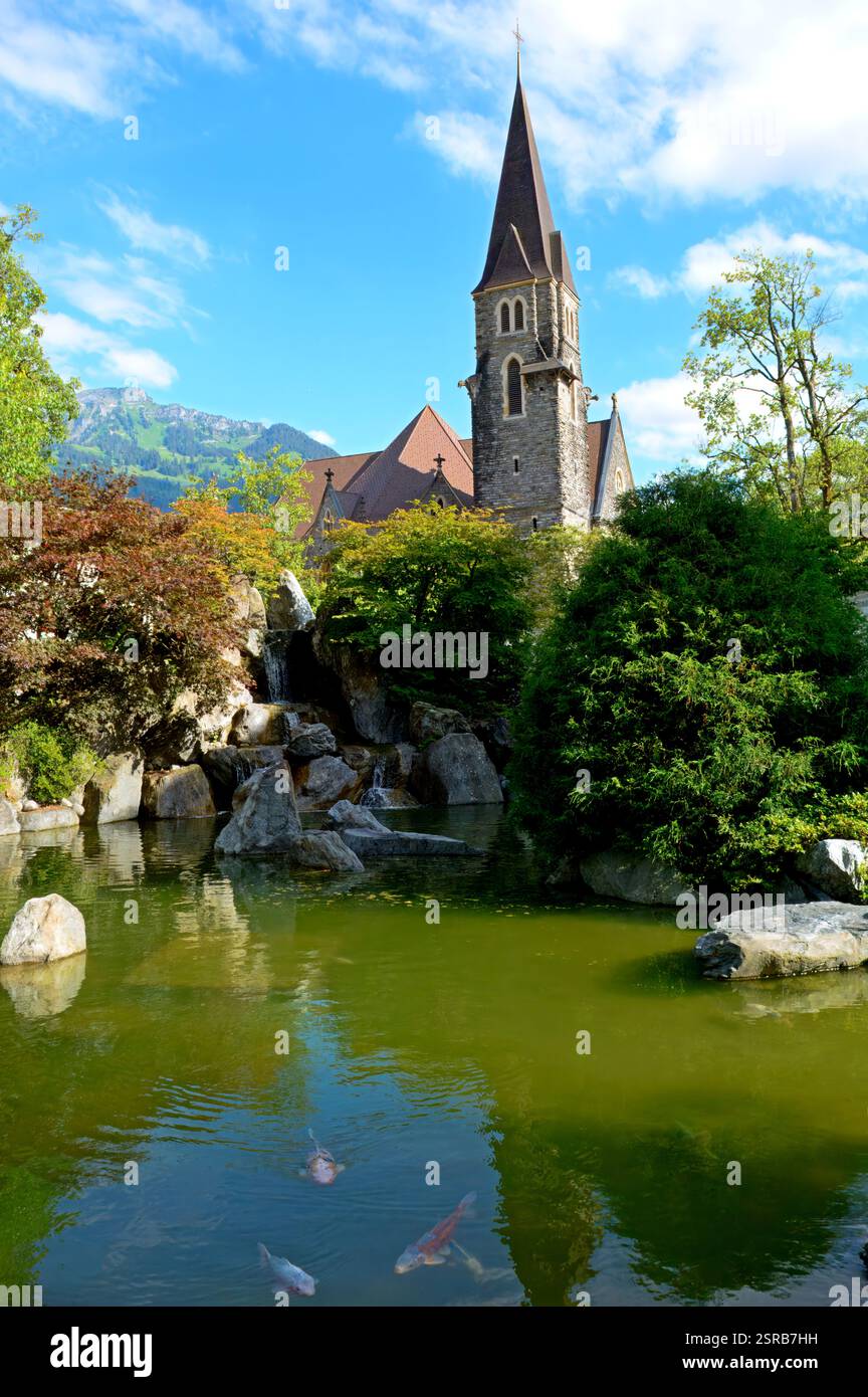 Una chiesa storica con un campanile a punta si affaccia su un tranquillo laghetto di koi con vegetazione lussureggiante, cascate e riflessi nell'acqua. Foto Stock