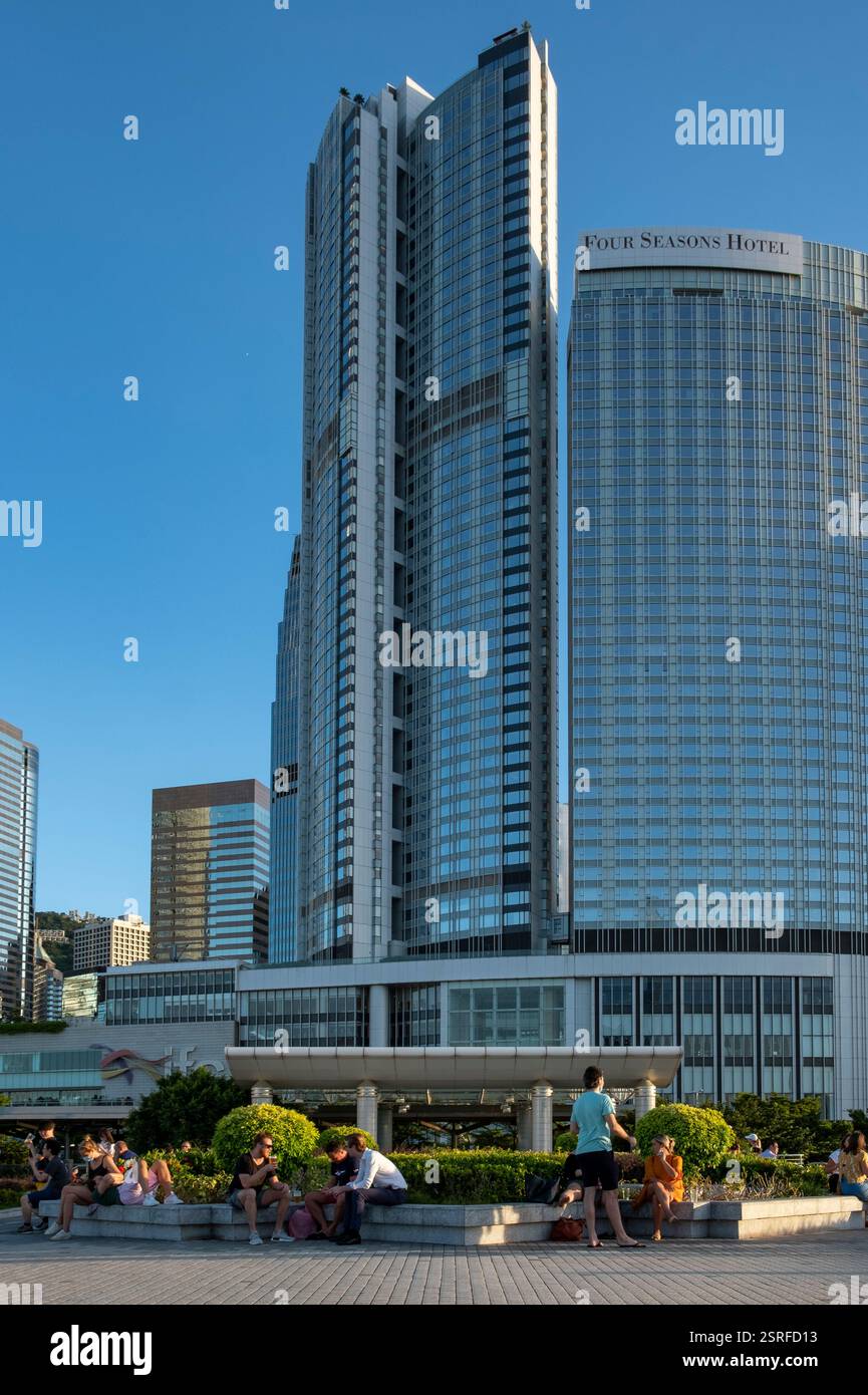 I giovani si godono un drink al Molo 4 nel centro di Hong Kong, con il Four Seasons Hotel e grattacieli sullo sfondo in un pomeriggio di sole. Foto Stock