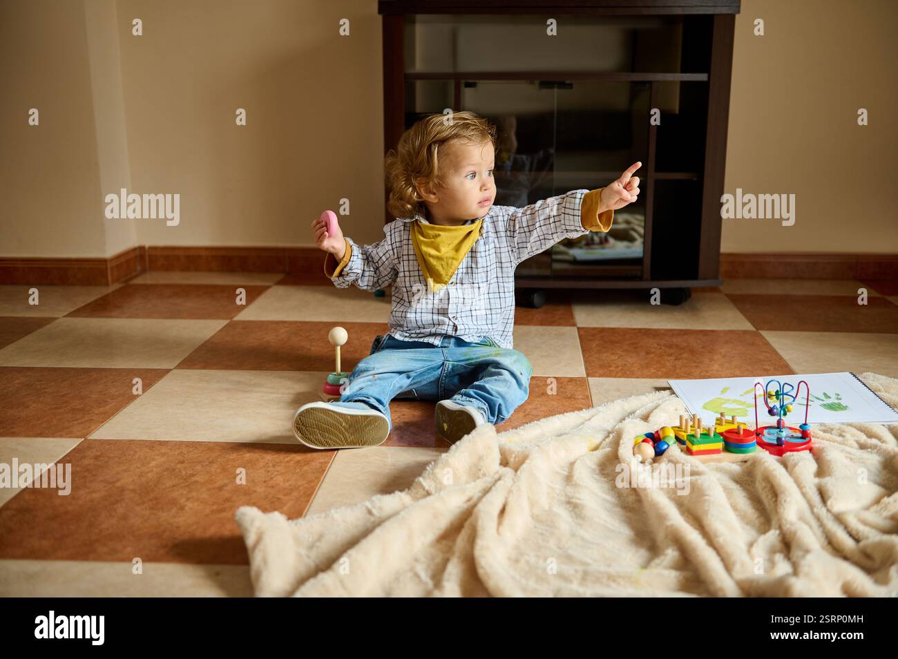 Un bambino siede sul pavimento in interni circondato da giocattoli, esprimendo curiosità e gioia mentre punta verso qualcosa con un look mirato. Foto Stock