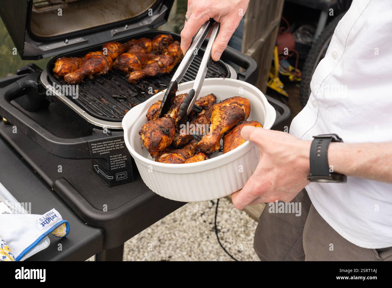 Uomo che rimuove le cosce di pollo cotte alla griglia da un forno all'aperto (Ninja Woodfire) con pinze in una ciotola, Regno Unito. Concetto: Cucina all'aperto Foto Stock