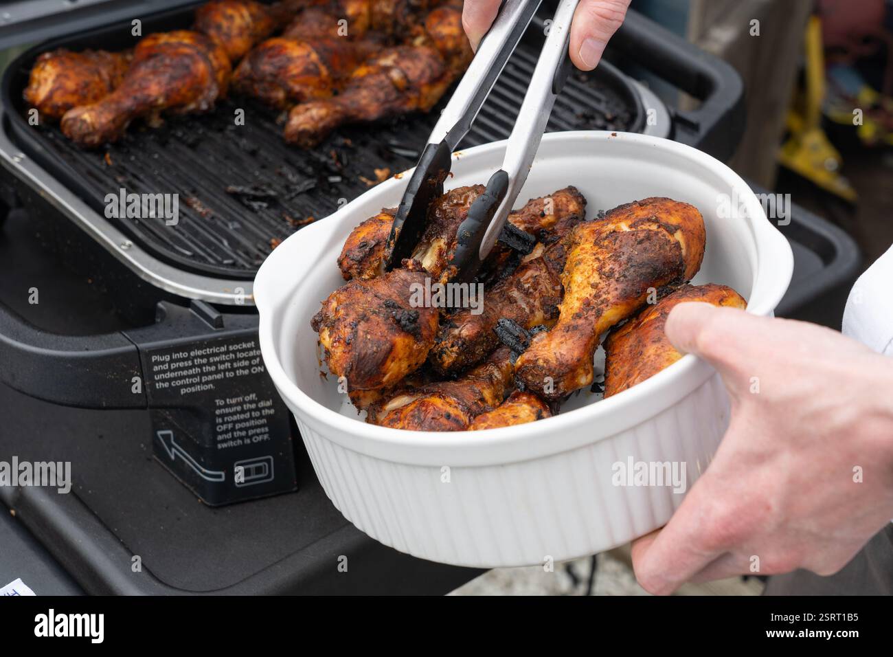 Uomo che rimuove le cosce di pollo cotte alla griglia da un forno all'aperto (Ninja Woodfire) con pinze in una ciotola, Regno Unito. Concetto: Cucina all'aperto Foto Stock