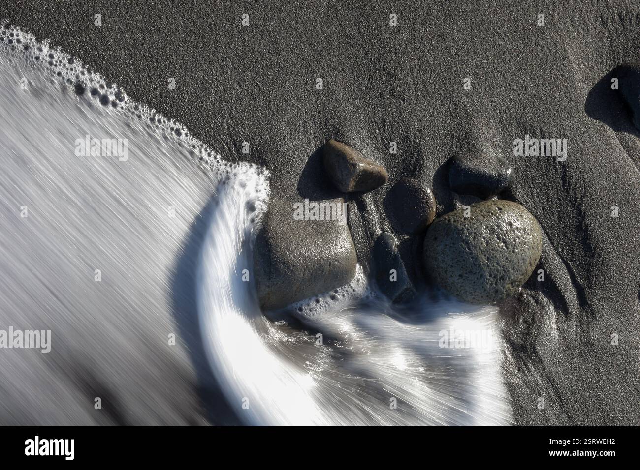 Una lenta velocità dell'otturatore cattura le onde dell'oceano che scorrono su sabbia nera e pietre lisce su una spiaggia tranquilla Foto Stock