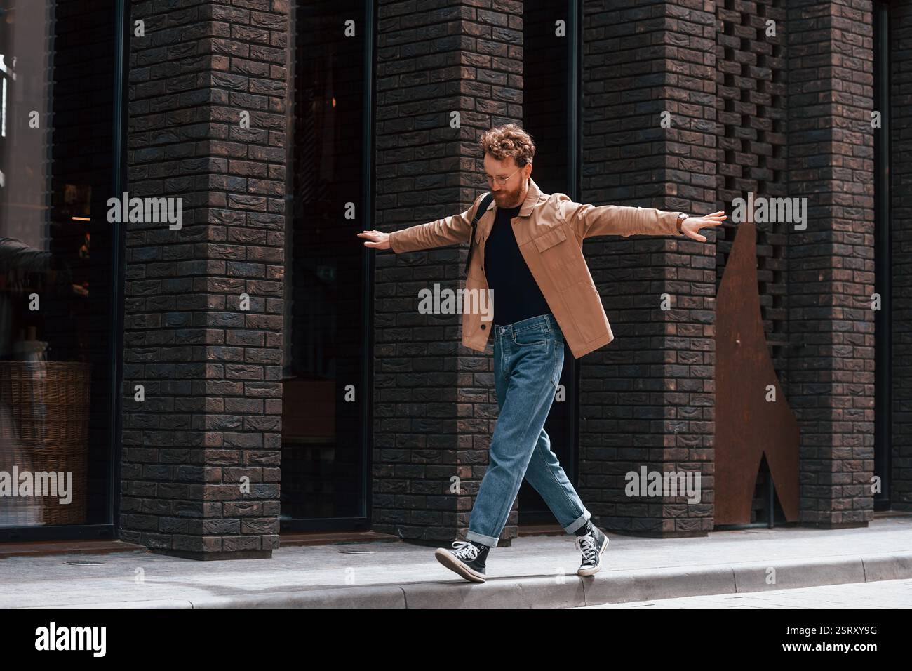 Divertitevi camminando sul marciapiede. Un uomo elegante con barba in giacca color cachi e jeans si trova all'aperto vicino all'edificio. Foto Stock
