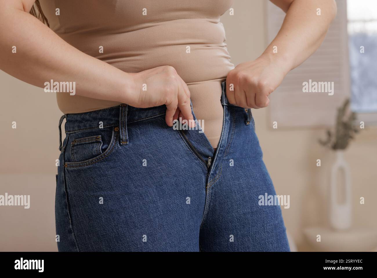 Una donna lotta per indossare i jeans stretti, evidenziando l'aumento di peso, i cambiamenti del corpo, le sfide di moda e la positività del corpo in un momento impareggiabile. Foto Stock