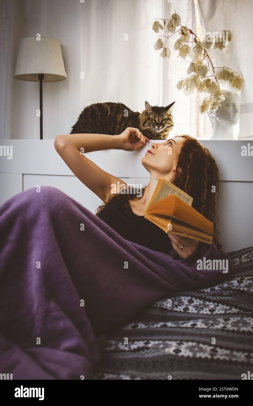 La donna si sta rilassando a casa. Sdraiato a letto con un libro e un gatto Foto Stock