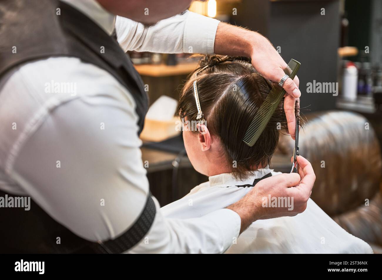 Vista posteriore del barbiere in un elegante abito che taglia i capelli bagnati. Giovane cliente che ottiene un taglio di capelli elegante. Un parrucchiere professionale ti offre un'acconciatura fresca. Foto Stock