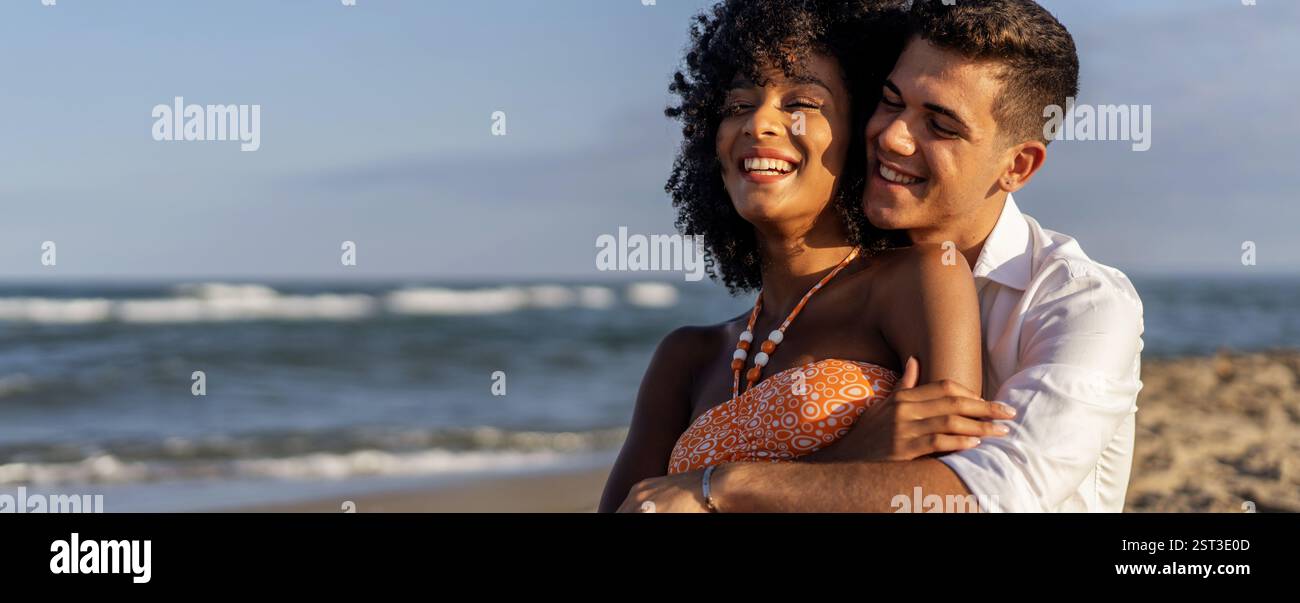 Allegra giovane coppia interrazziale che abbraccia la spiaggia, ridendo e godendosi una romantica vacanza estiva sull'oceano. Concetto di amore, libertà e. Foto Stock