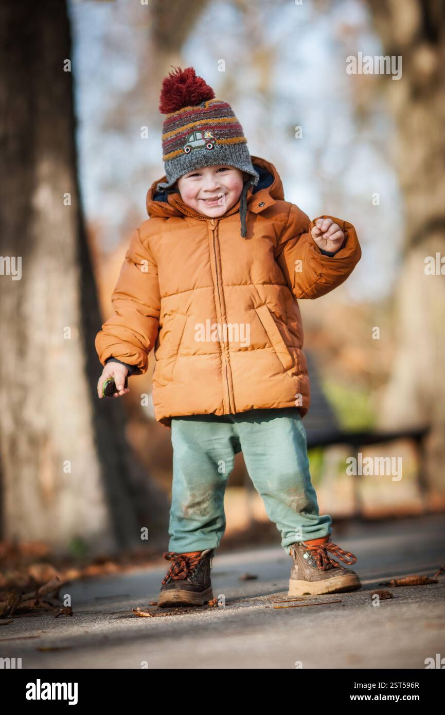 Un bambino che indossa un giubbotto di peluche arancione e un cappello a maglia sorride mentre gioca in un parco in autunno. Le foglie circostanti mostrano una vivace caduta Foto Stock