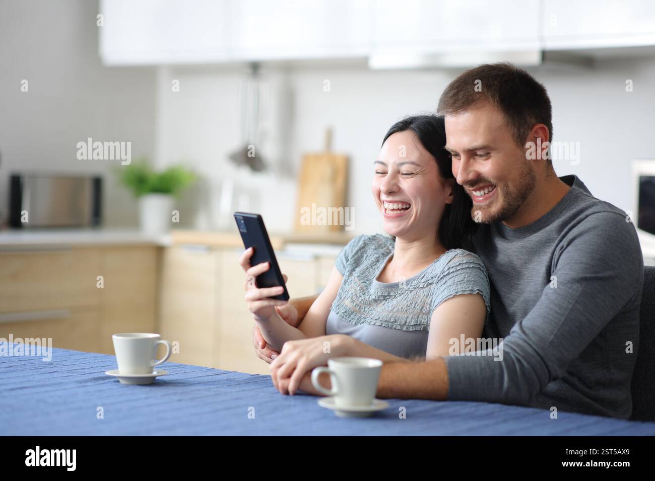 Felice coppia interrazziale che ride controllando lo smartphone in cucina a casa Foto Stock
