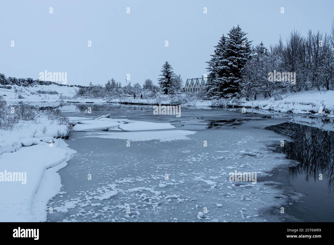 Un tranquillo paesaggio invernale dove il fiume ghiacciato riflette le sfumature morbide del paesaggio innevato. Case nordiche tradizionali sullo sfondo. Foto Stock