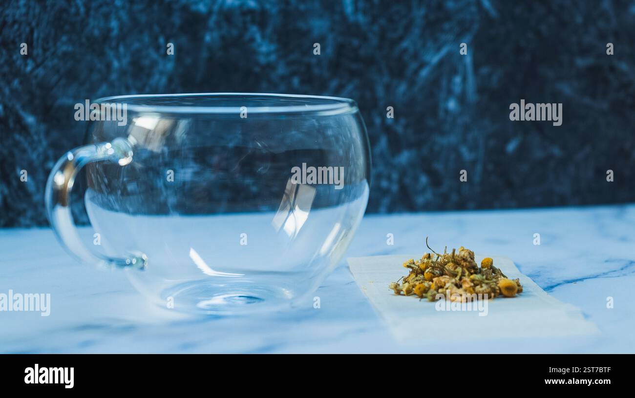 Tazza di vetro accanto ai fiori di camomilla secchi su una superficie di marmo in un ambiente accogliente da cucina Foto Stock