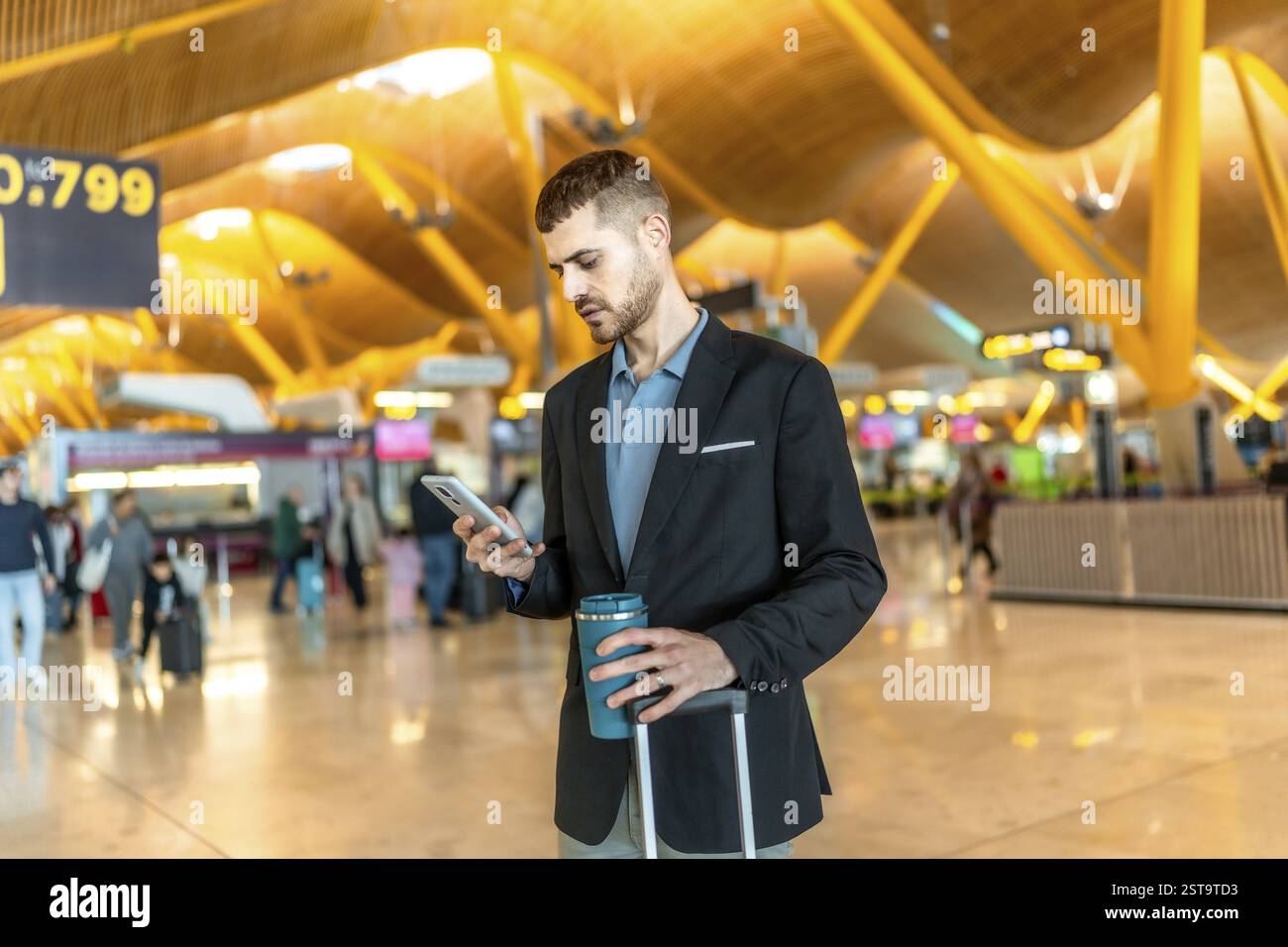 Uomo d'affari in attesa del volo, che usa lo smartphone e tiene in mano una tazza di caffè riutilizzabile all'aeroporto Foto Stock