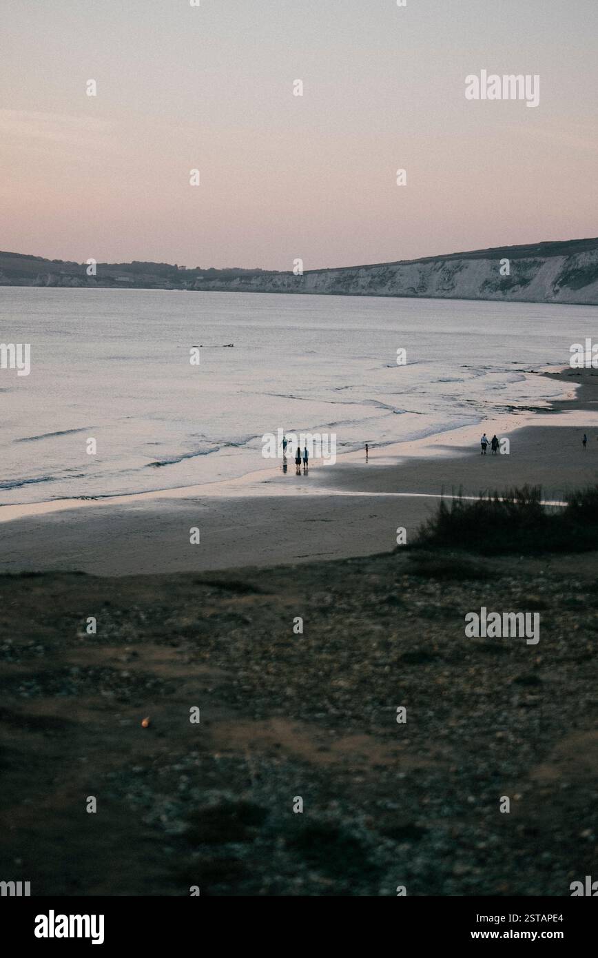 Un gruppo di persone sta camminando su una spiaggia al tramonto. Il cielo è un mix di tonalità arancio e rosa, che creano un'atmosfera serena e tranquilla. La spiaggia lo è Foto Stock