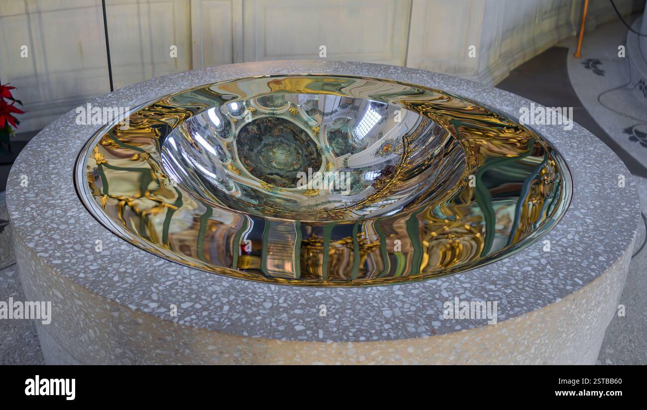 Sankt Gallen, Svizzera - 1° gennaio 2025: Moderno fonte battesimale con un bacino metallico riflettente situato in una base di pietra in una cattedrale di Sankt Gall Foto Stock