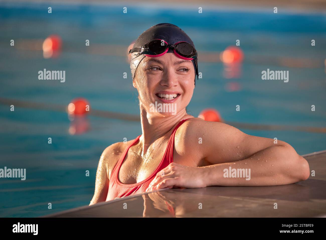 Un nuotatore in costume rosso sorride sul bordo della piscina, rivestito di occhiali protettivi, dopo una sessione di allenamento concentrata in una struttura di nuoto al coperto Foto Stock