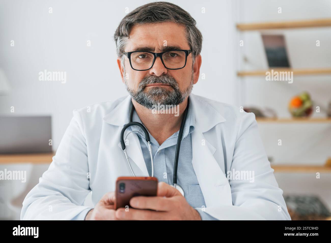 Un medico maschile è seduto con il telefono in mano. Operatore medico professionale in cappotto bianco è in ufficio. Foto Stock