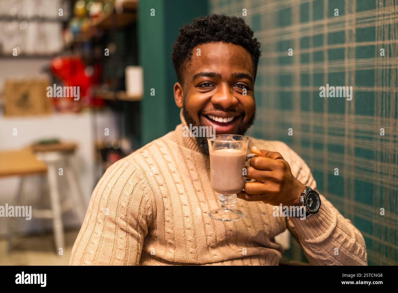 Giovane uomo nero allegro che beve cioccolata calda al bar. Ritratto di un uomo che tiene la cioccolata calda, guarda la macchina fotografica e sorride. Foto Stock