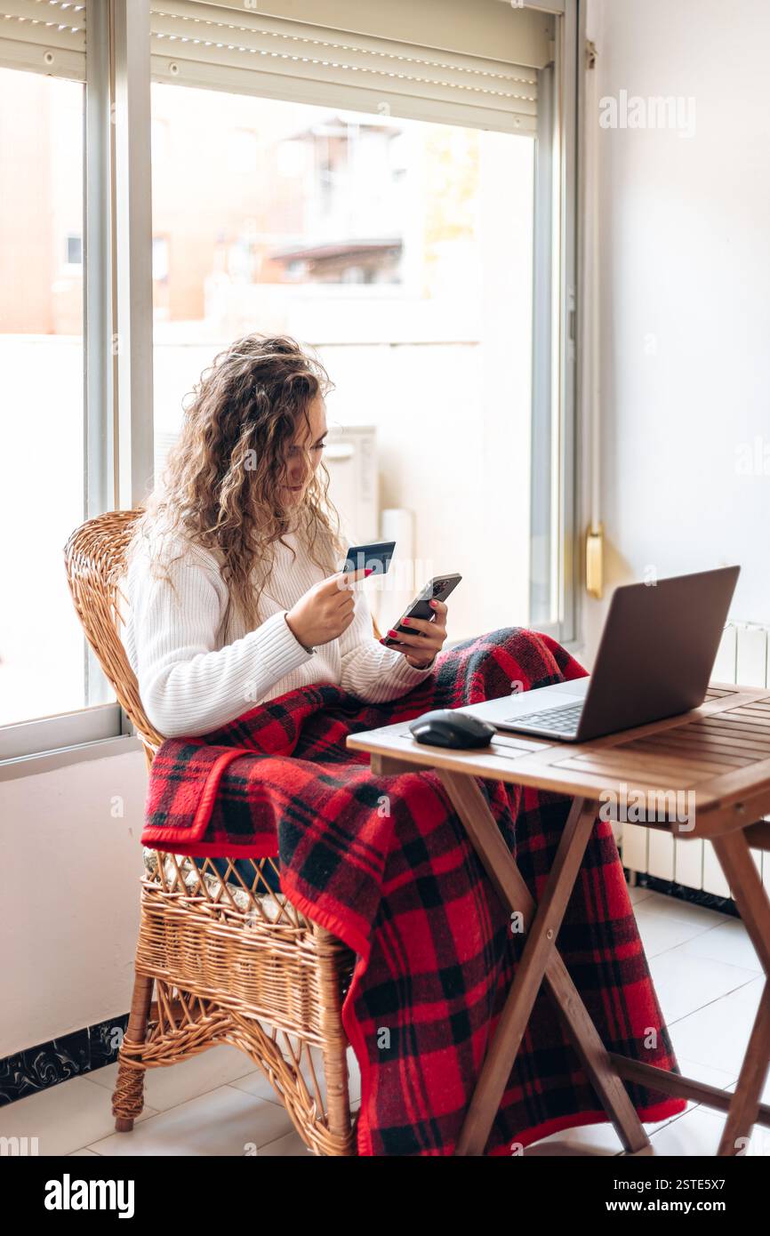 Giovane donna dai capelli ricci in maglione bianco seduto a tavola con il laptop mentre utilizza lo smartphone e la carta di credito per l'acquisto online via Internet Foto Stock
