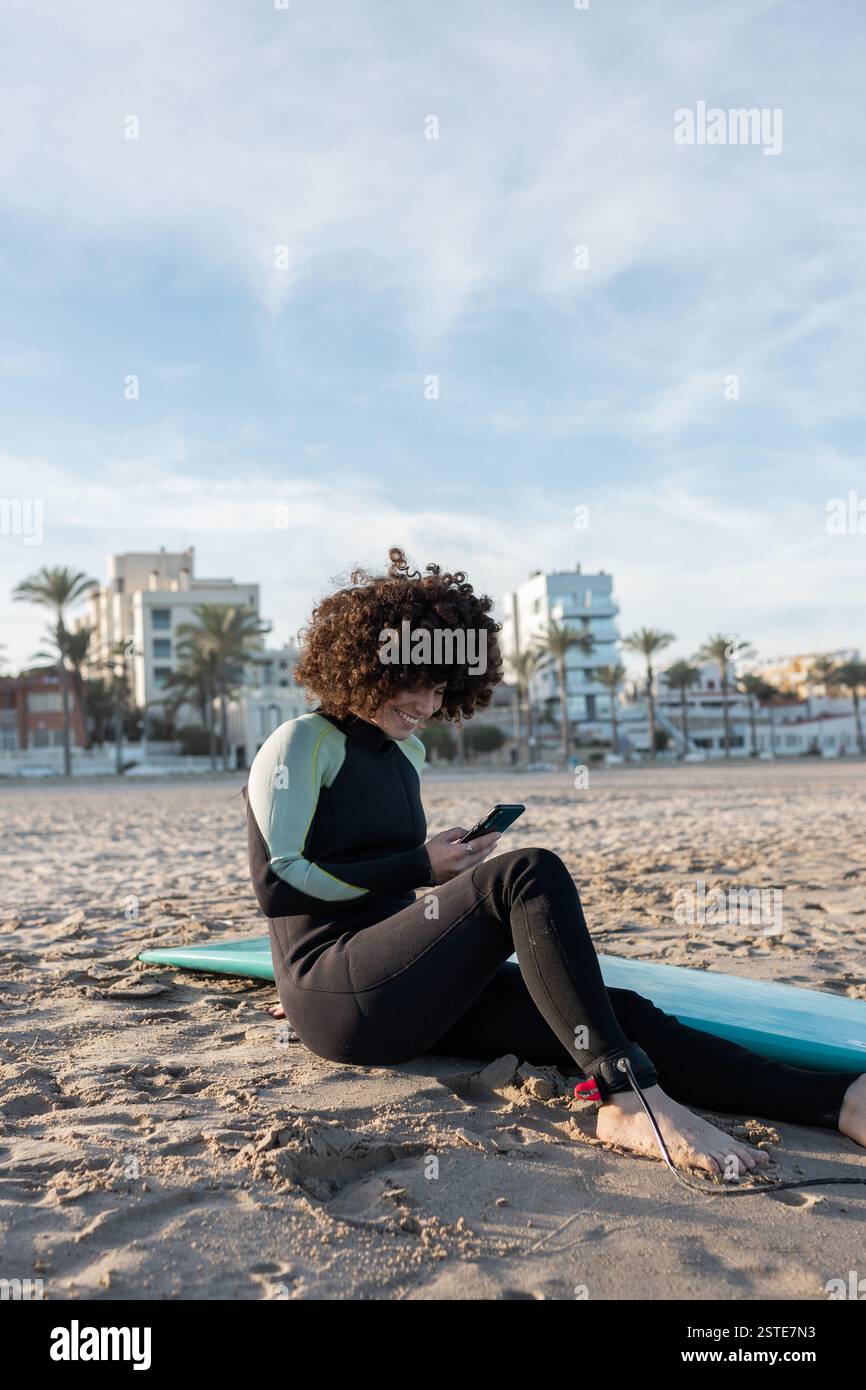 Donna positiva con capelli ricci indossando la muta che naviga smartphone mentre si siede su una spiaggia sabbiosa con tavola da surf Foto Stock