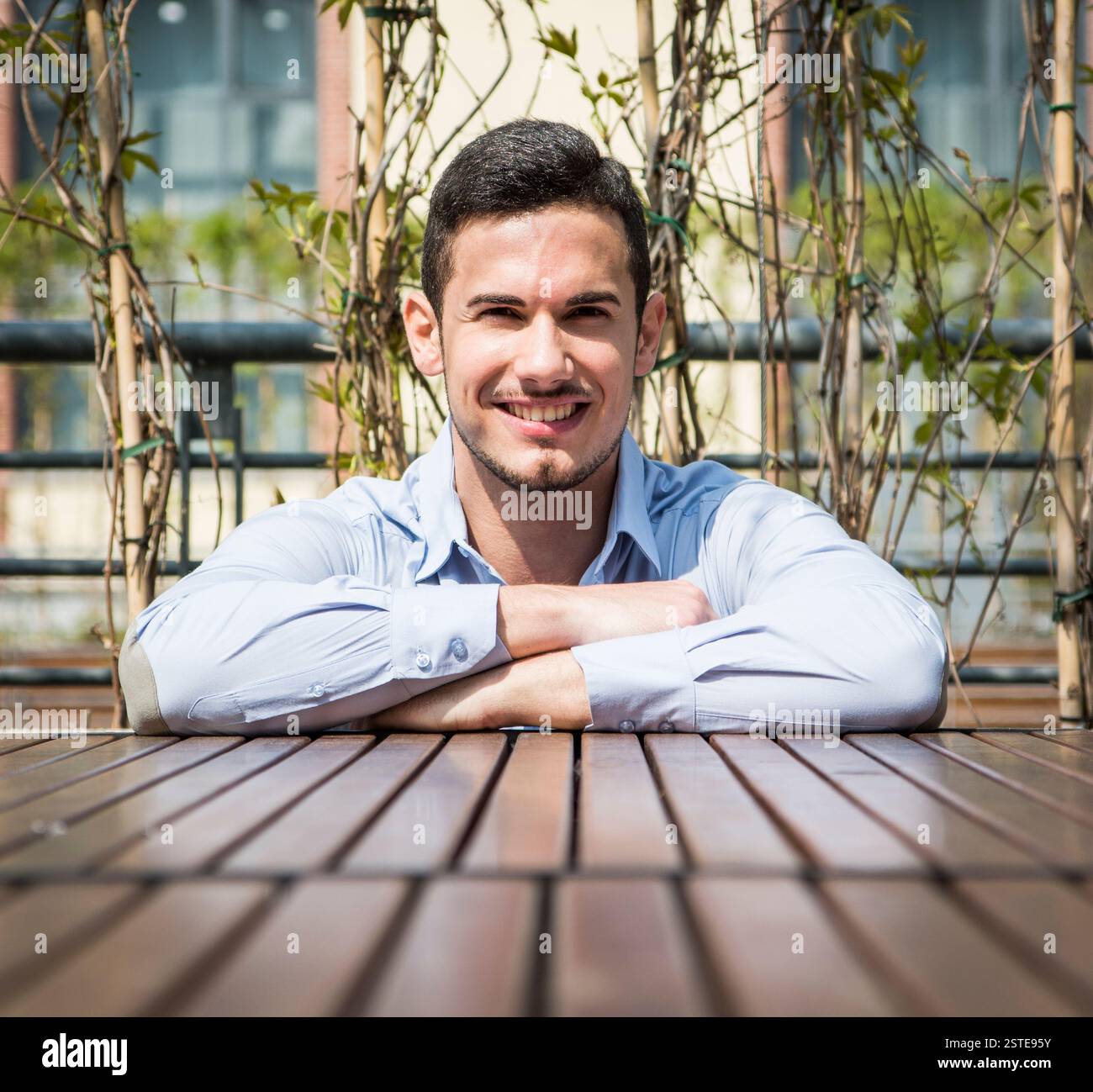 Bel giovane uomo appoggiato bracci incrociati sulla superficie di legno mentre cercano e sorridente in telecamera Foto Stock