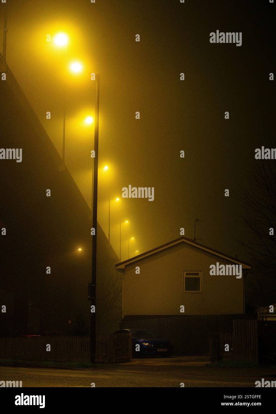Una casa proprio sotto un cavalcavia autostradale in una notte nebbiosa con i semafori sul ponte che formano una linea diagonale Foto Stock