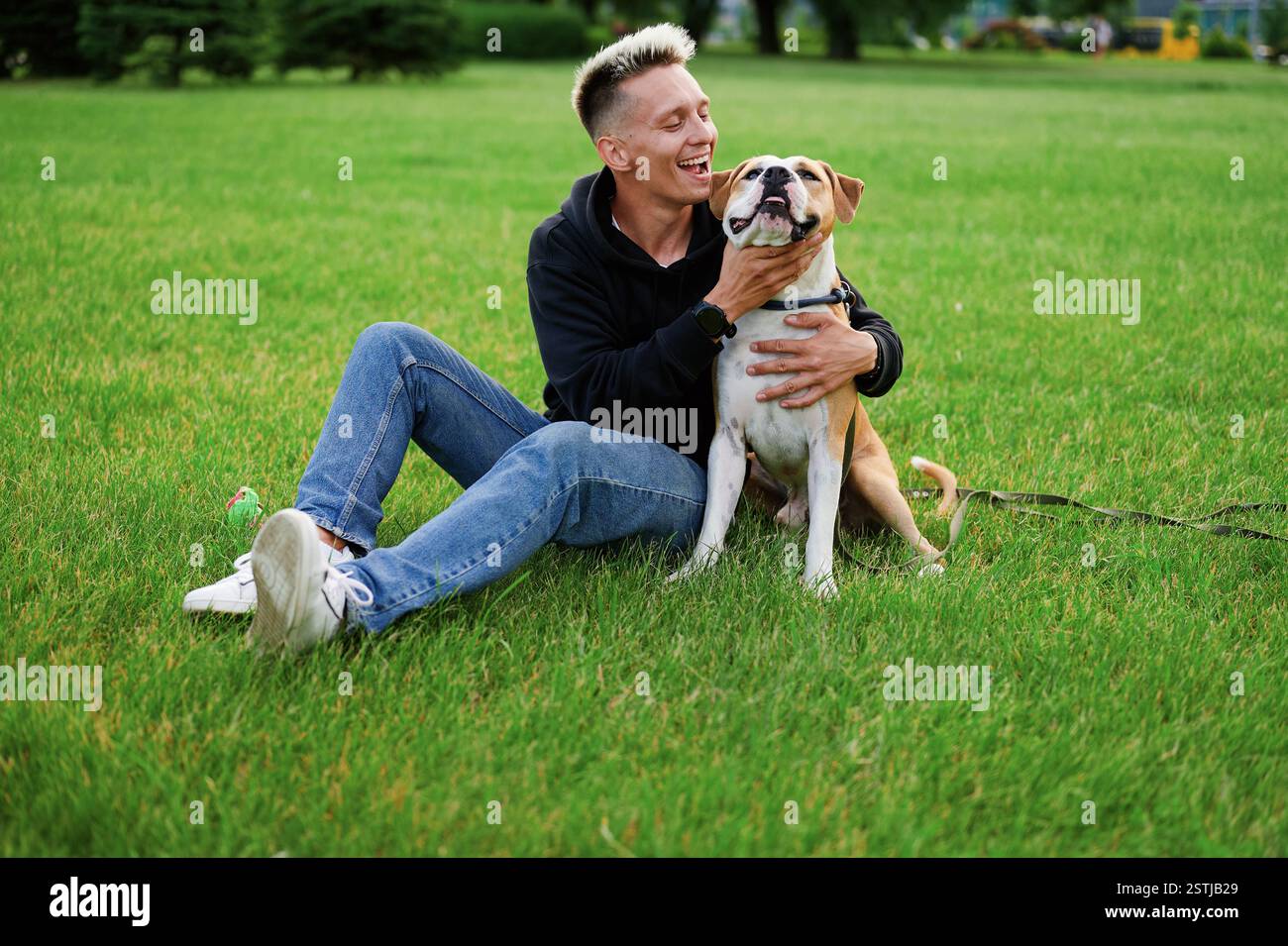 Guy e il suo cane, bulldog americano, natura. Uomo felice e il suo cane all'aperto in estate. Il giovane uomo caucasico coccola il suo cane seduto sul gras Foto Stock