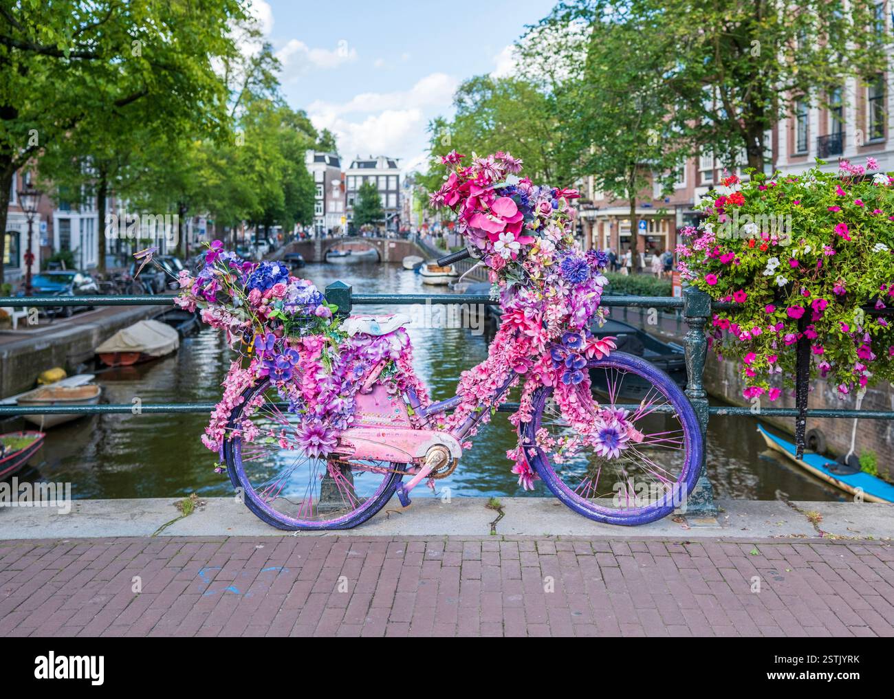 Una bicicletta decorata in modo luminoso ricoperta di fiori rosa e viola poggia su un ponte sul canale di Amsterdam. Circondato da vegetazione lussureggiante Foto Stock