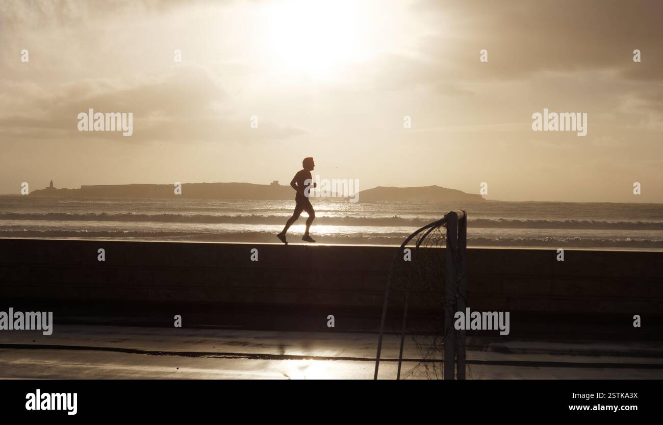 Jogger corre sul muro in silhouette dopo la pioggia con gol in primo piano e mare e isola alle spalle a Essaouira, in Marocco. 18 febbraio 2025 Foto Stock