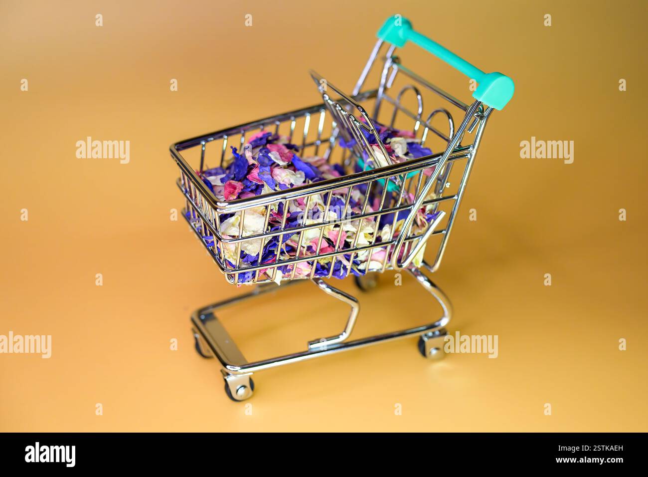 Un mini carrello della spesa contenente petali di fiori misti colorati in contrasto con uno sfondo liscio di colore giallo Foto Stock