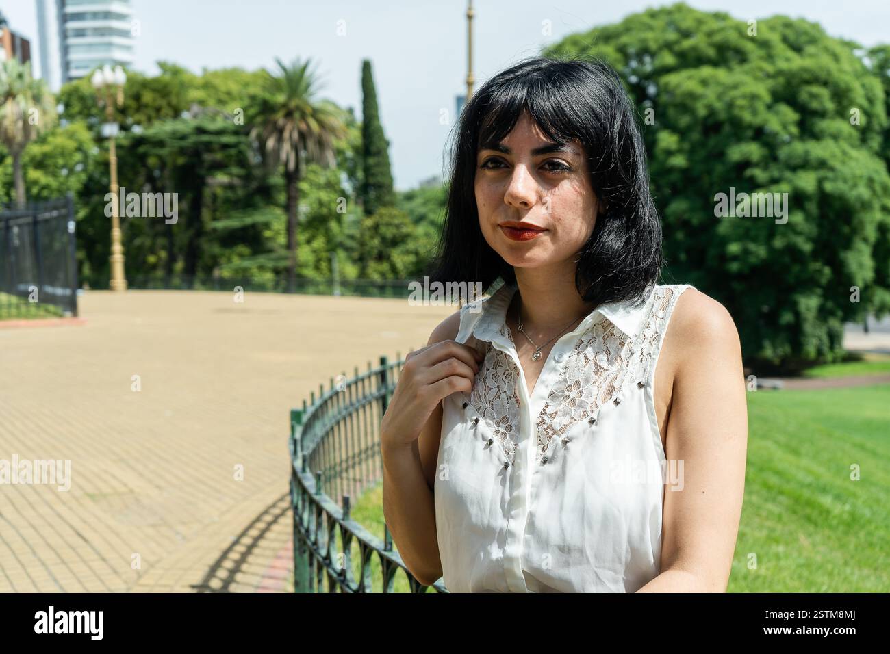 Elegante donna latina che regola la sua camicetta bianca mentre si trova vicino a una recinzione in un parco Foto Stock