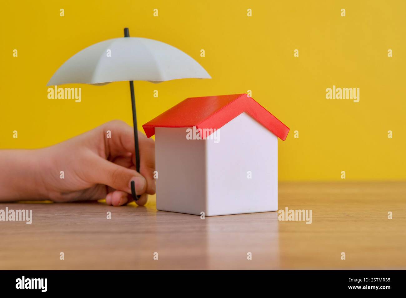 Una casa dei giocattoli è protetta da un ombrello in miniatura tenuto da una mano. Concetto di assicurazione per la casa. Foto Stock