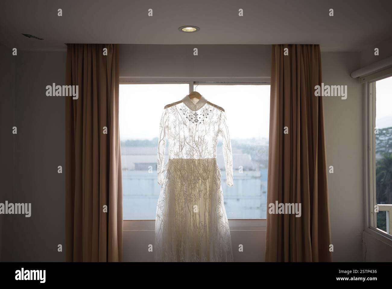Un elegante abito da sposa bianco appeso su un appendiabiti vicino alla finestra con tende marrone chiaro all'interno, che simboleggia la preparazione nuziale e un bellissimo c Foto Stock