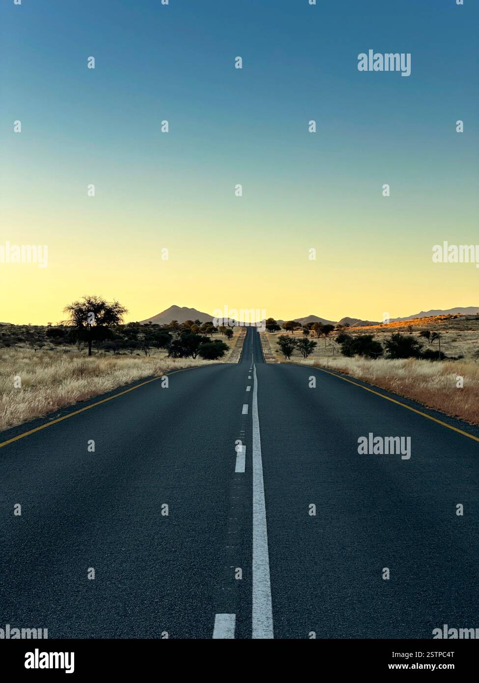 Una lunga strada rettilinea (C15) si estende all'orizzonte al tramonto, circondata da praterie asciutte e montagne lontane verso Dordabis, Namibia. - Immagine stock catturata con smartphone