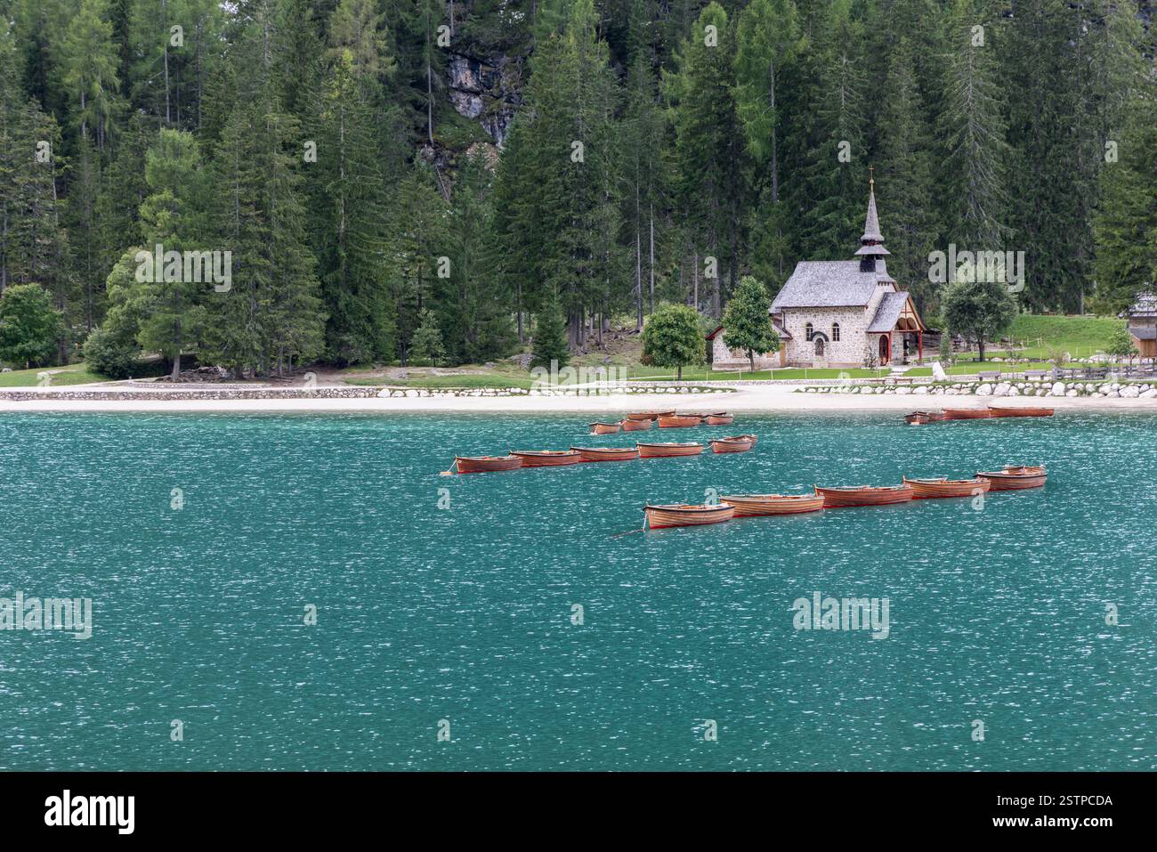 Una fila di barche in legno che galleggiano sulle acque turchesi vicino alla spiaggia sabbiosa del lago Braies con un'affascinante cappella sul lago e una fitta foresta sempreverde Foto Stock