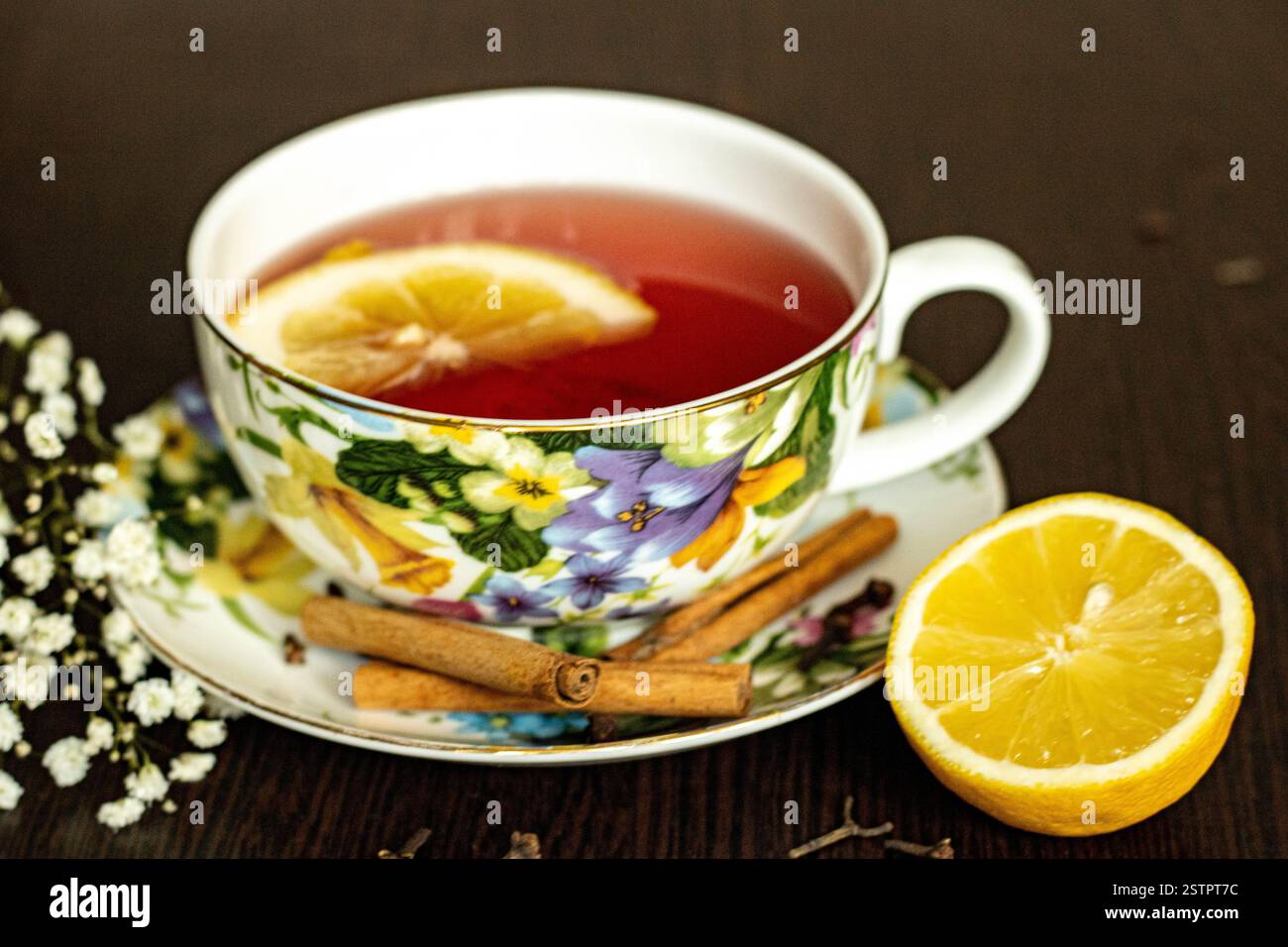 Tè rosso aromatico, con una fetta di limone, in una tazza molto ben decorata, su un simpatico piattino, con mezzo limone, piccoli fiori bianchi come decorazione Foto Stock