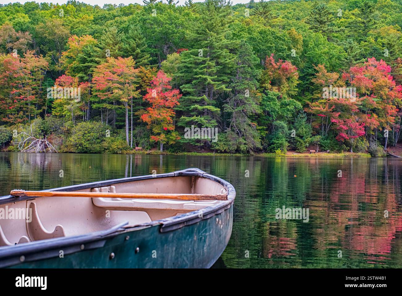 Una canoa rustica si trova sulle acque calme di un lago, riflettendo i vibranti alberi autunnali in tonalità rosse, arancioni e verdi lungo la costa. Foto Stock