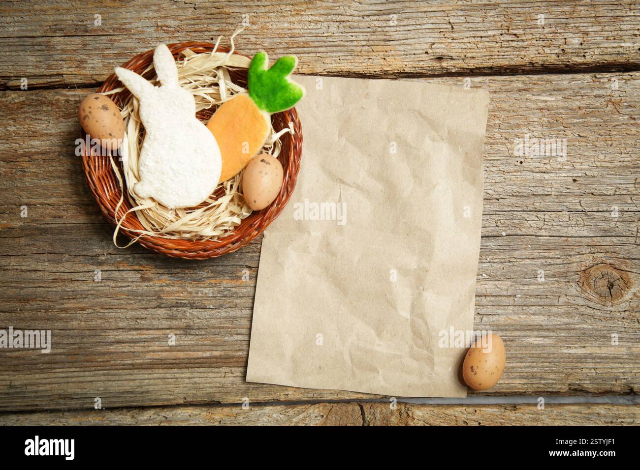 Vista sul tavolo dei biscotti di zucchero e delle uova di Pasqua in un nido su fondo rustico in legno con carta bianca, Mock Up per biglietti d'auguri o fornelli Foto Stock