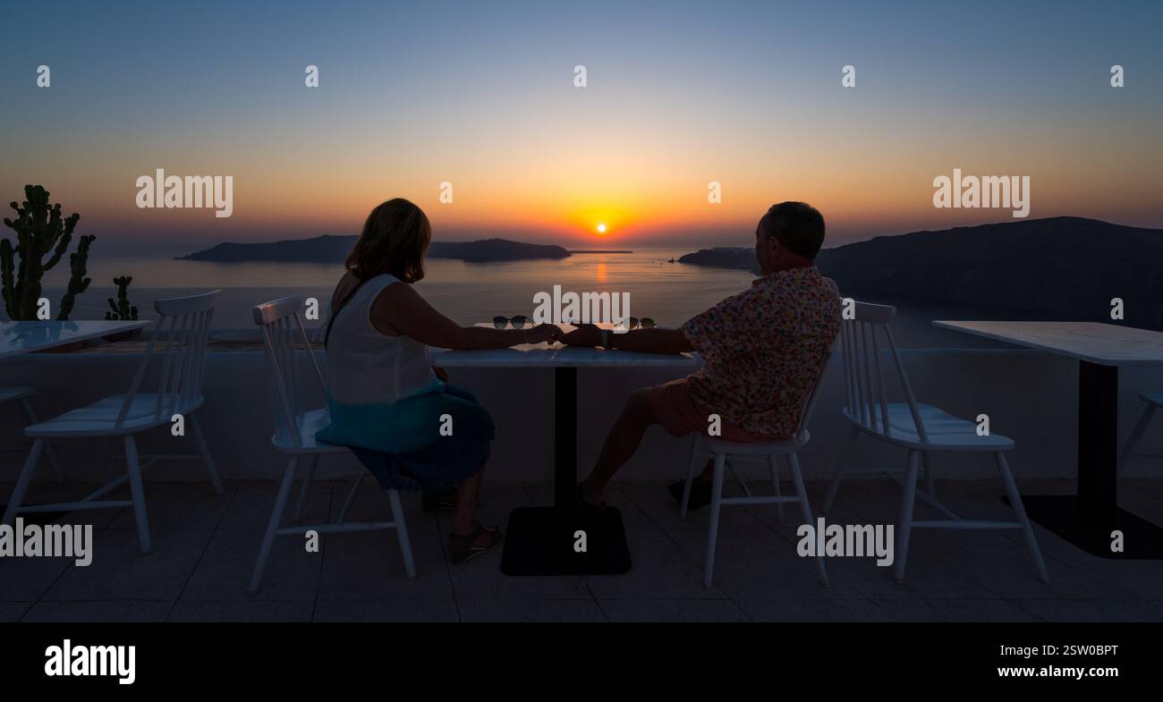 La coppia sedeva per mano al tavolo e si affacciava sul tramonto drammatico sulla caldera di Santorini Foto Stock