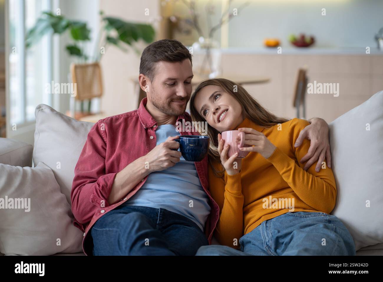 Coppia felice che si abbraccia sul divano, beve caffè, tè. Coniugi soddisfatti che si rilassano in un caldo abbraccio. Foto Stock