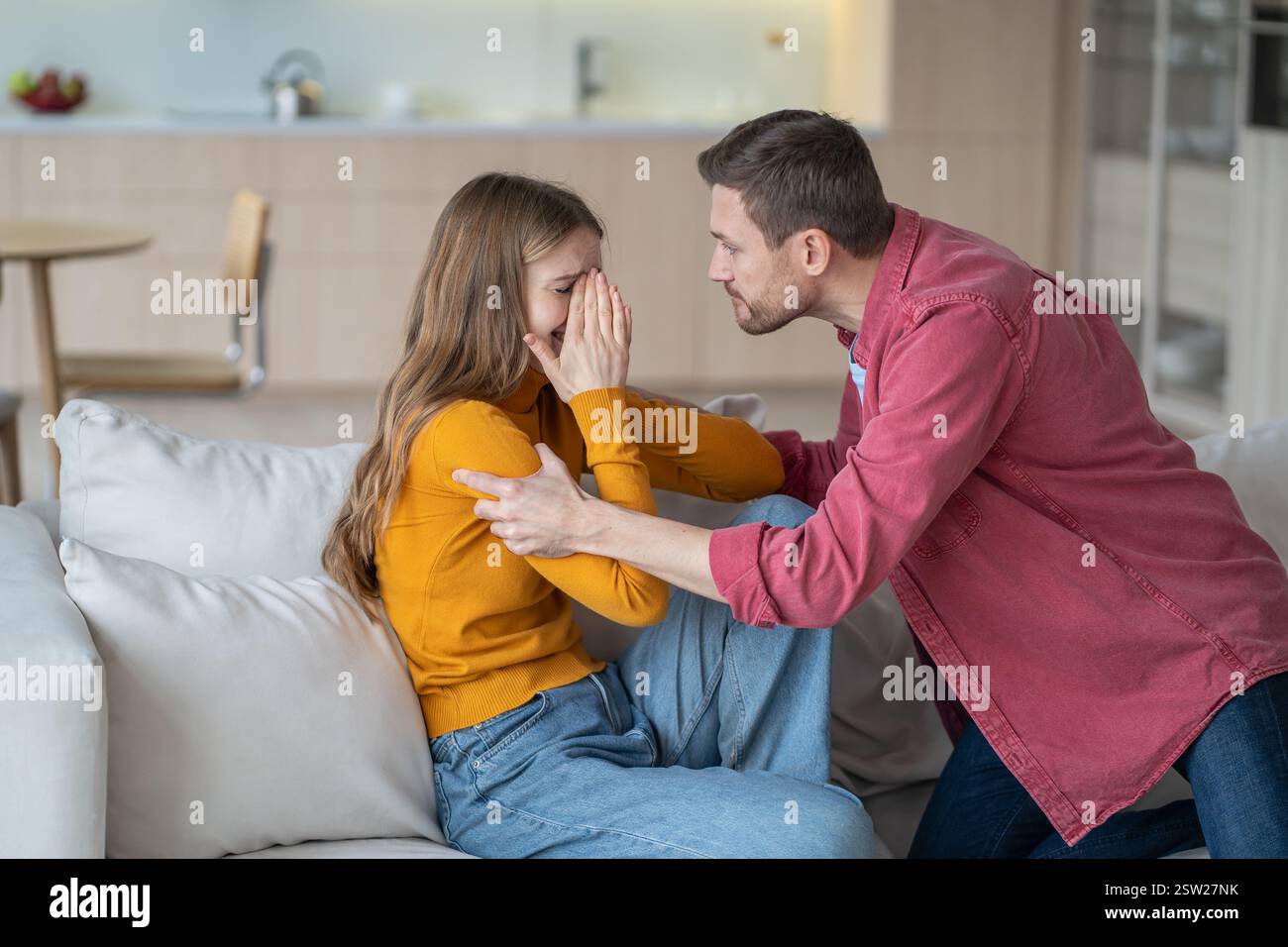 Il marito tiranno grida e scuote piangendo la moglie temuta, litigando e afferrando le mani sul divano Foto Stock