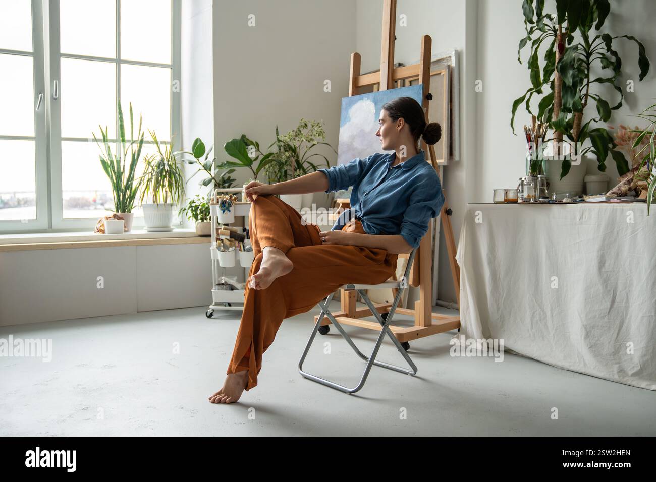 Una donna espressiva e premurosa artista seduto in posa a gambe incrociate guarda fuori dalla finestra nello studio d'arte creativo Foto Stock
