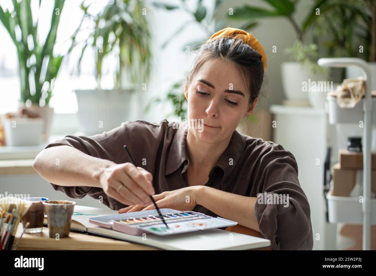 Artista donna concentrata che crea con acquerelli e pennello nell'album si siede sul pavimento in uno studio d'arte. Foto Stock
