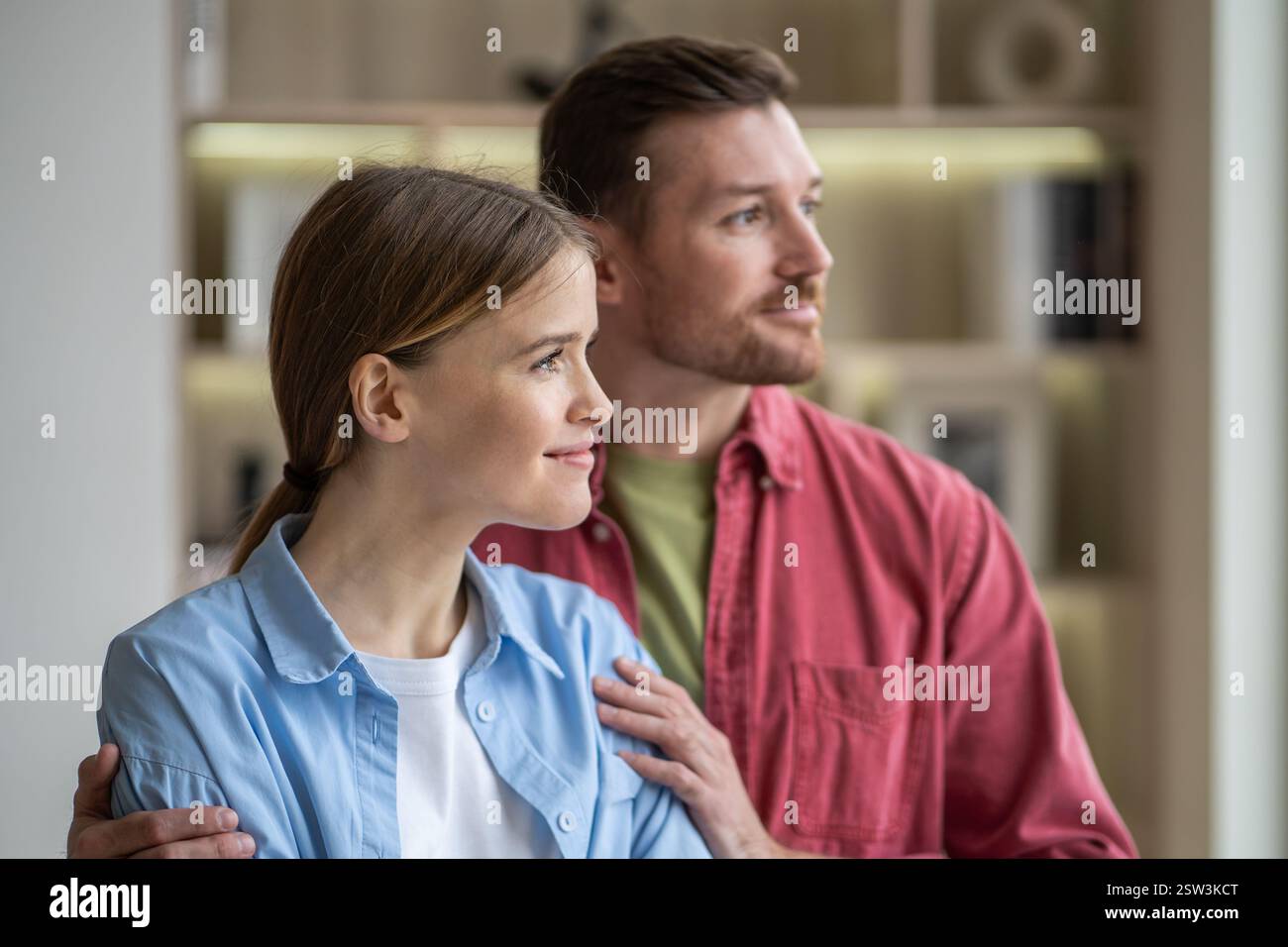 Una coppia romantica e serena che abbraccia e guarda finestra in un accogliente appartamento. Conforta il matrimonio, amore Foto Stock