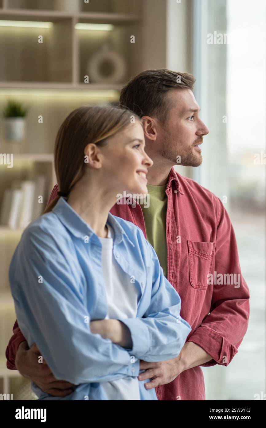 Gioiosa e sorridente, la giovane famiglia si abbraccia e guarda fuori dalla finestra, nella casa accogliente. Legame di coppia, intimità a casa Foto Stock