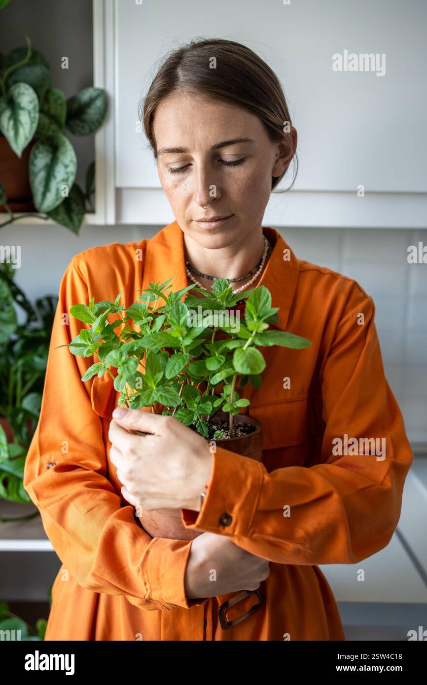 Una donna contenta e tranquilla abbraccia un vaso di menta profumata coltivata nel giardino di casa su davanzale. Amante delle piante Foto Stock