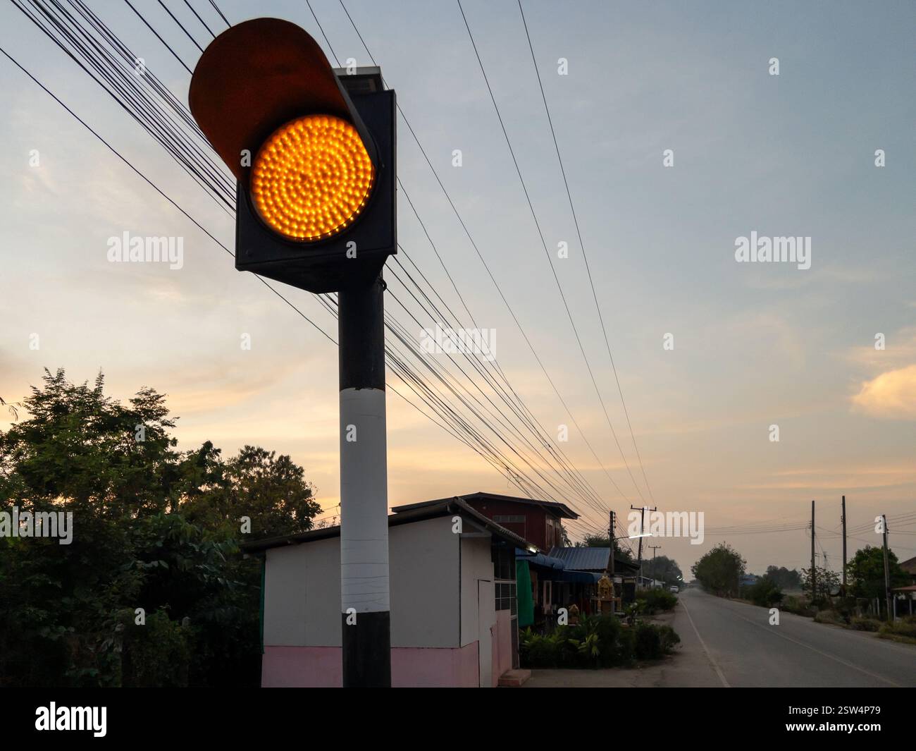 Un semaforo a LED color ambra si erge in alto in corrispondenza di un incrocio rurale e si illumina in modo luminoso contro la luce del giorno che sbiadisce. Le linee elettriche si estendono lungo la S Foto Stock