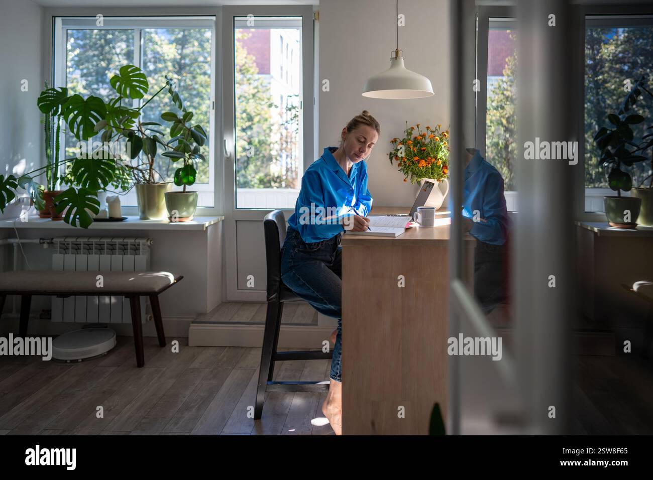 Una donna concentrata e confortevole lavora a casa scrivendo appunti nell'agenda giornaliera, seduto a un tavolo con un laptop. Foto Stock