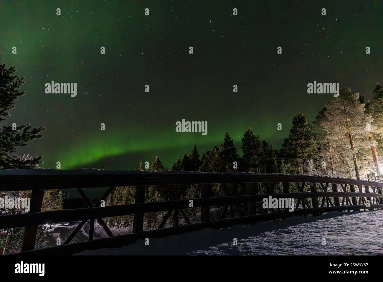 un brillante arco verde dell'aurora boreale è basso nel cielo sopra il parapetto di un ponte innevato in una foresta della lapponia finlandese Foto Stock