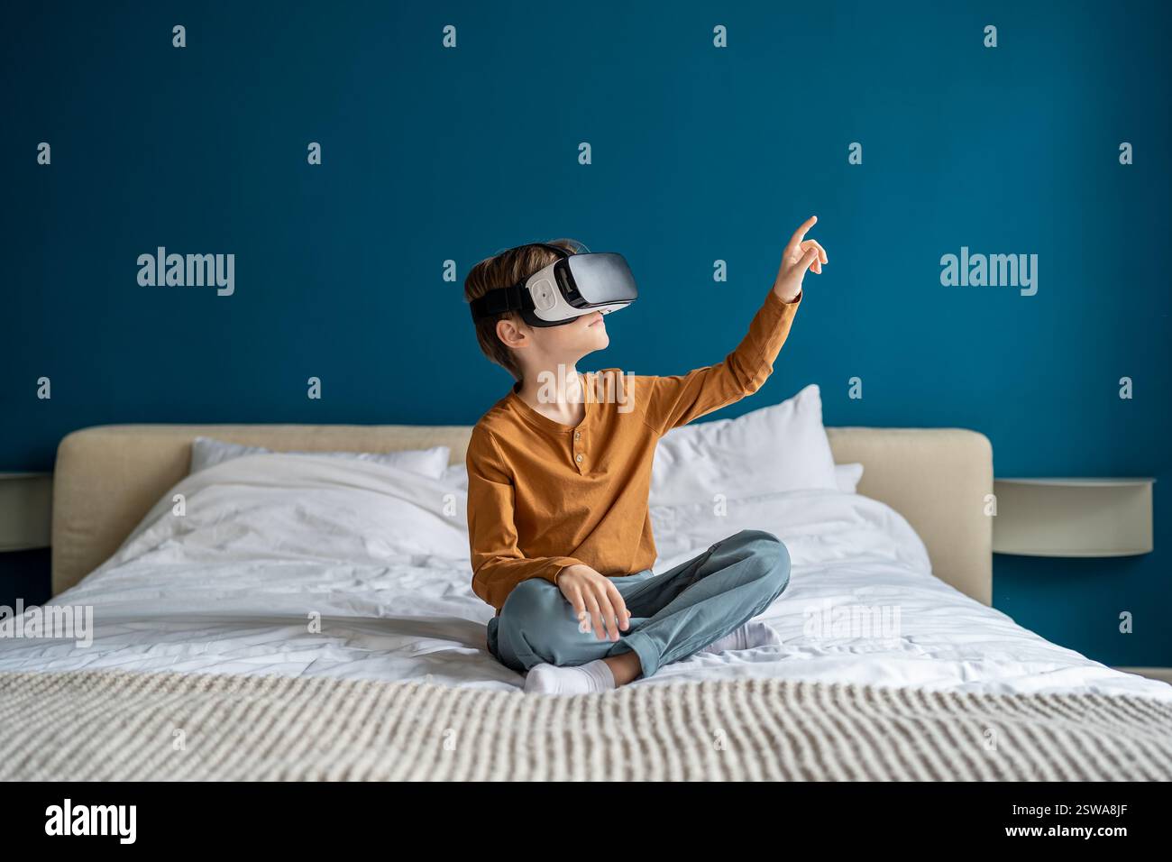 Bambino che indossa un visore per la realtà virtuale seduto sul letto di casa a esplorare la metaversa. Concetto di realtà virtuale Foto Stock